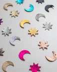 Flat lay of assorted crescent moon and star shapes made from colourful, textured leather in metallic and bold finishes, arranged in a scattered pattern on a light surface