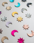 Flat lay of assorted crescent moon and star shapes made from colourful, textured leather in metallic and bold finishes, arranged in a scattered pattern on a light surface