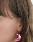 Close-up of a woman wearing a crescent moon-shaped earring in pink and white patterned leather with gold hook, contrasting softly with her natural hair tones