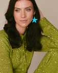Model wearing a sequinned lime green outfit paired with a bright turquoise blue star-shaped leather earring, photographed in soft studio light