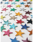 Flat lay of colourful leather star shapes arranged diagonally on a white background, featuring a mix of bold, metallic, and pastel tones