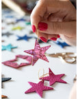 Hand holding a pink leather star earring over a table scattered with star-shaped leather pieces in various colours