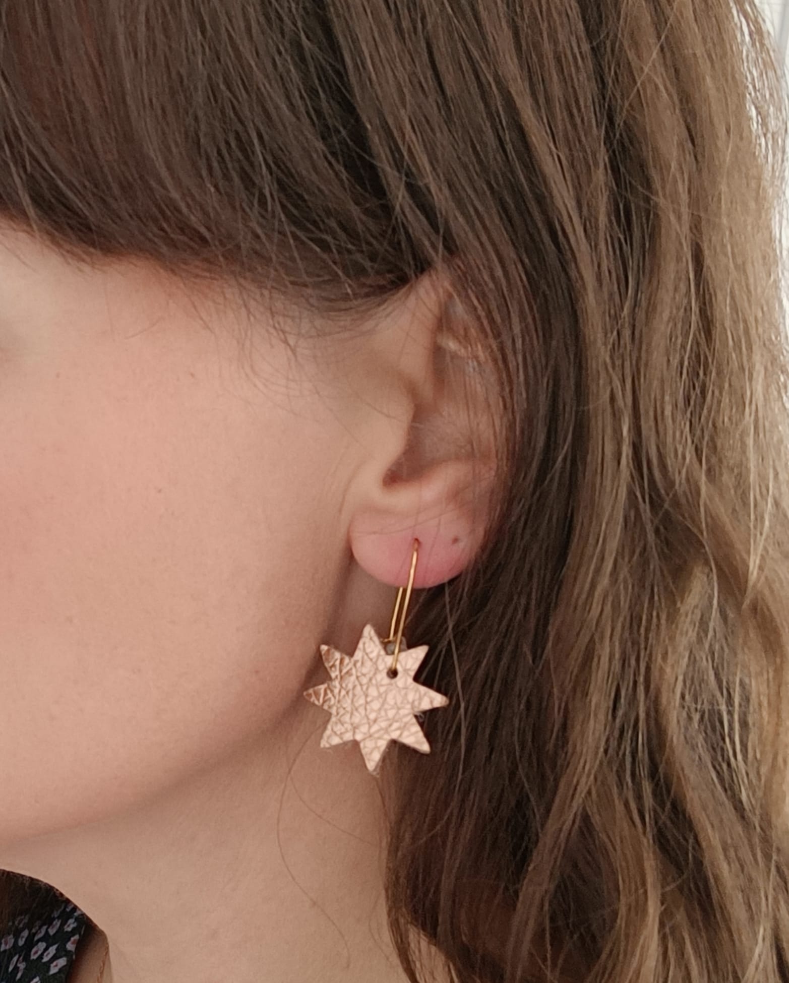 Close-up of a woman wearing a single rose gold leather star earring with gold hook, showing the earring's size and texture against her skin and hair