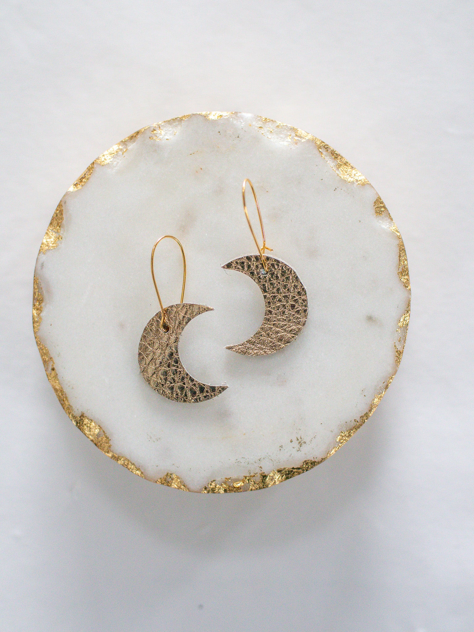 Gold leather crescent moon earrings with gold hooks, displayed on a white marble tray with gold edging