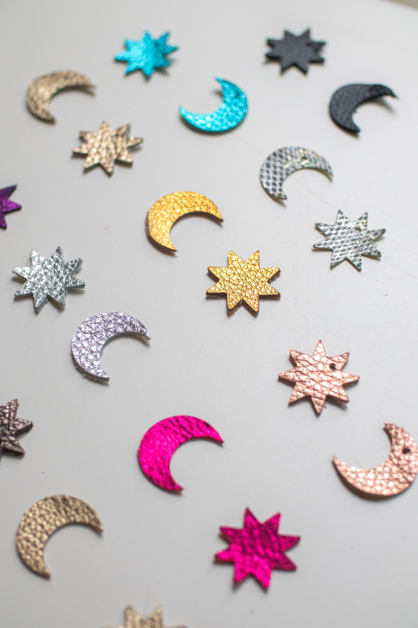 Flat lay of assorted crescent moon and star shapes made from colourful, textured leather in metallic and bold finishes, arranged in a scattered pattern on a light surface