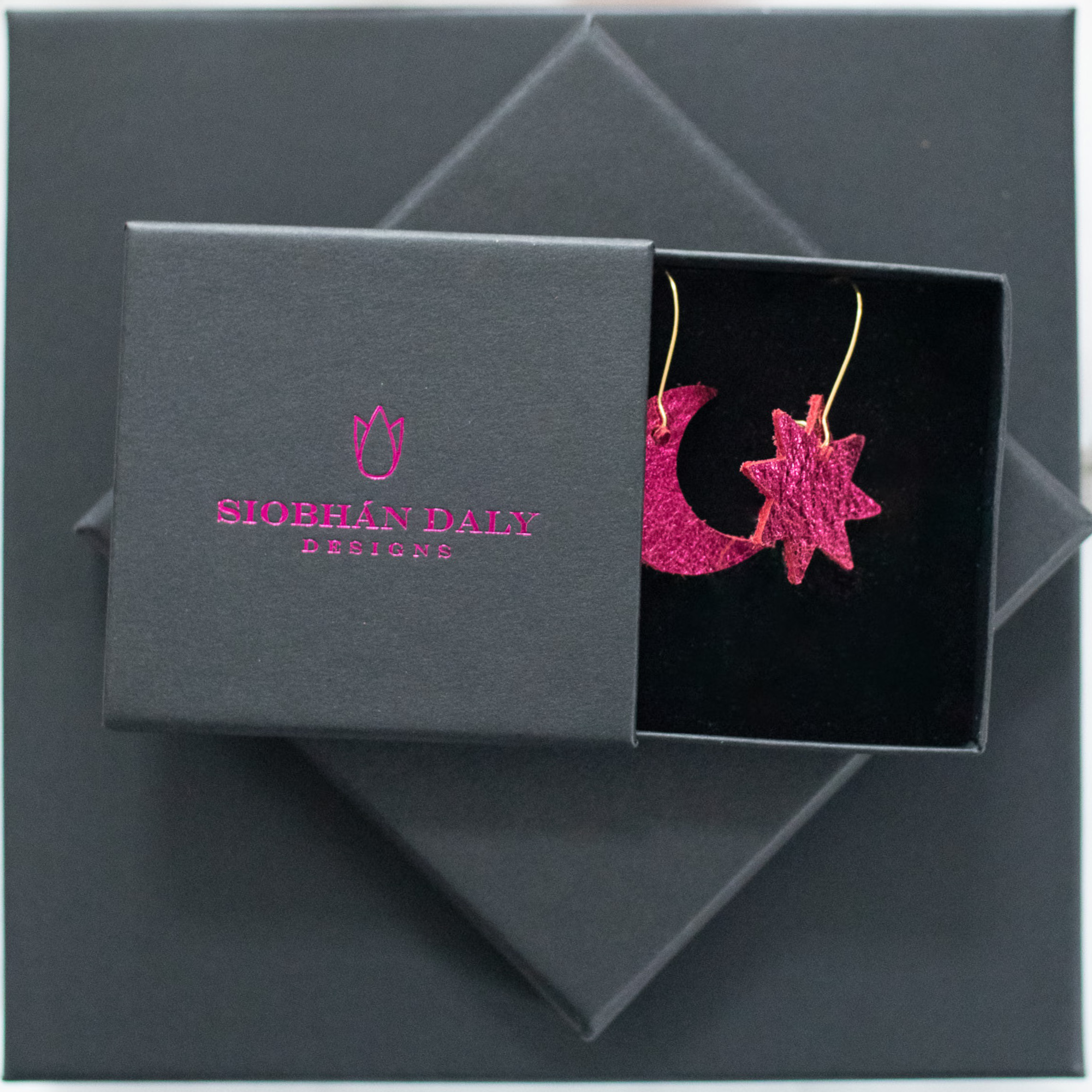 Metallic pink leather moon and star earrings with gold hooks, presented in a Siobhán Daly Designs branded gift box