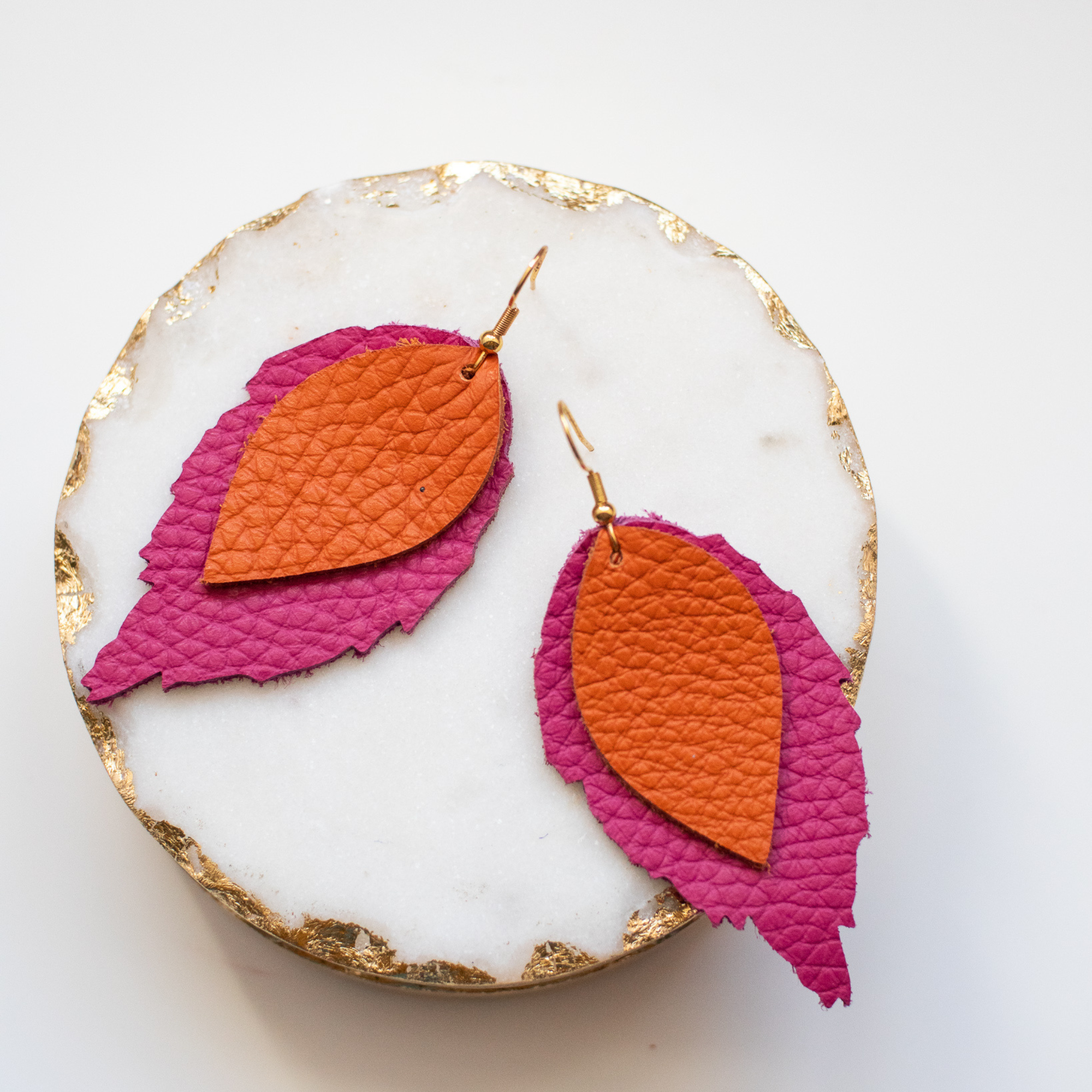 Layered leather earrings in orange and magenta pink, handcrafted from Italian top-grain leather and photographed on a white marble dish with gold trim