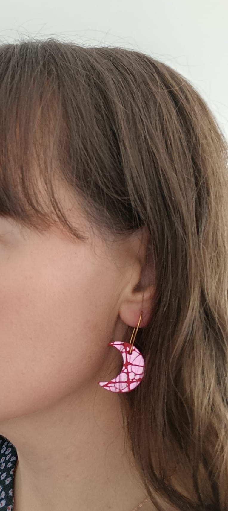 Close-up of a woman wearing a crescent moon-shaped earring in pink and white patterned leather with gold hook, contrasting softly with her natural hair tones
