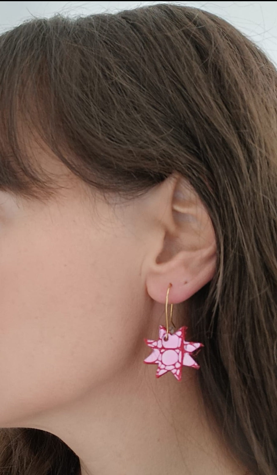 Close-up of a woman wearing a pink and white patterned leather star earring with gold hook, highlighting the bold colours and graphic design against her hair and skin