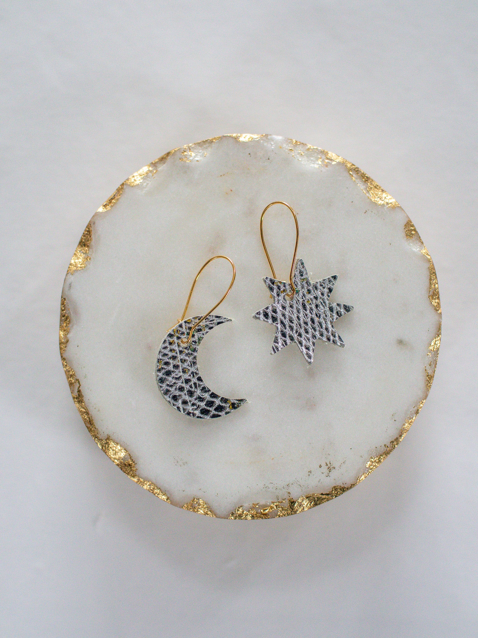 Silver leather moon and star earrings with gold hooks, displayed on a round white marble tray with gold trim