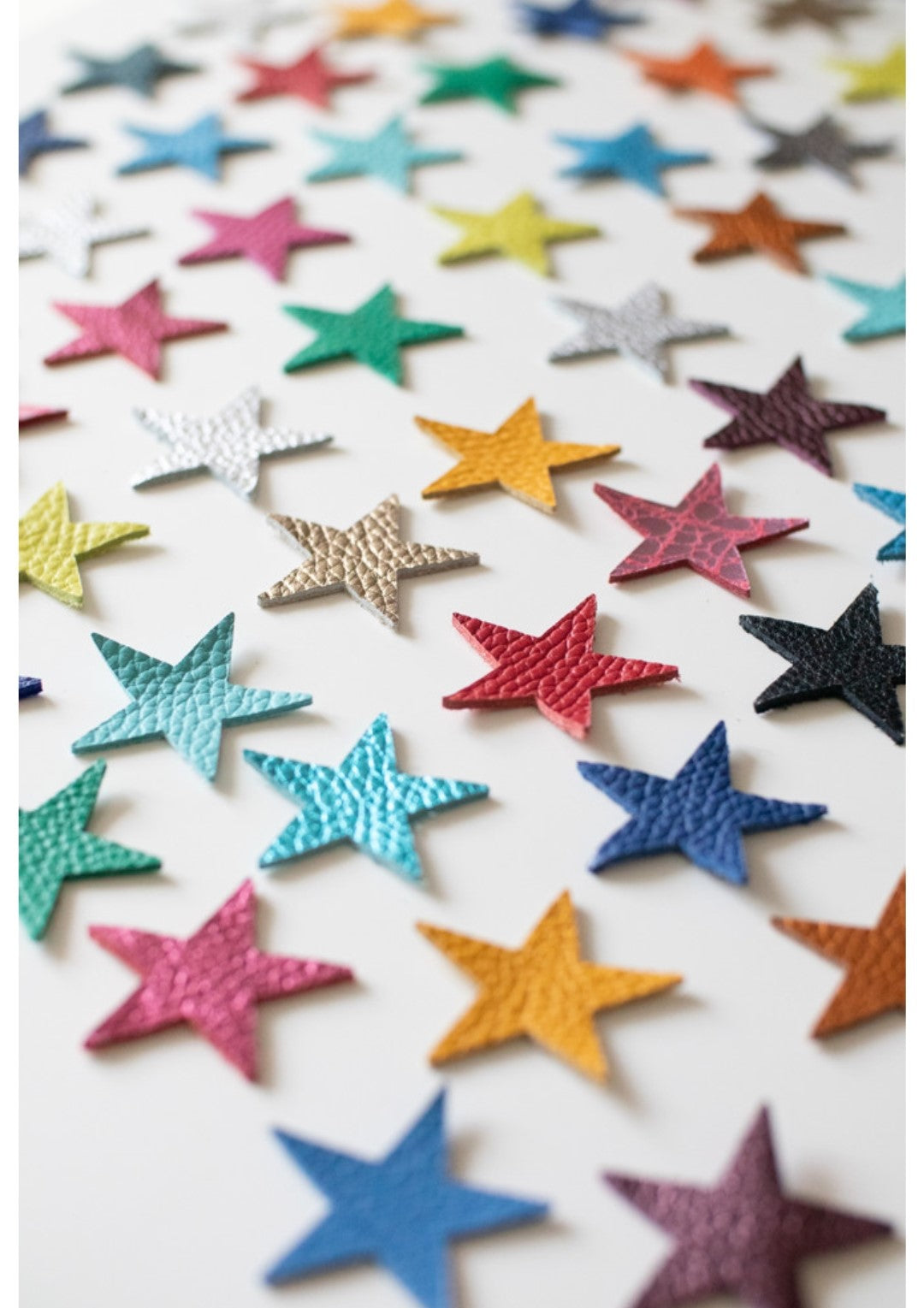Flat lay of colourful leather star shapes arranged diagonally on a white background, featuring a mix of bold, metallic, and pastel tones