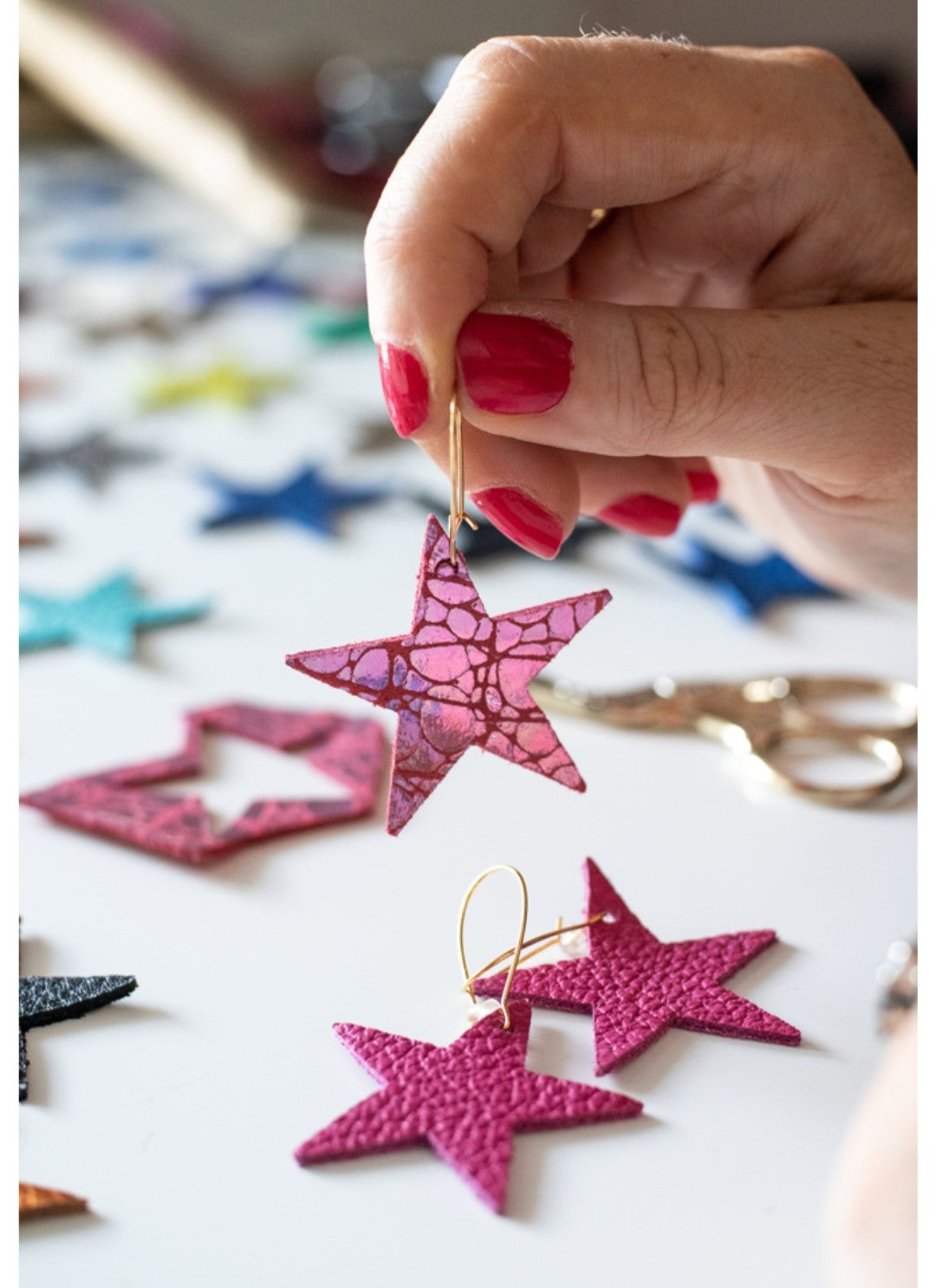 Hand holding a pink leather star earring over a table scattered with star-shaped leather pieces in various colours