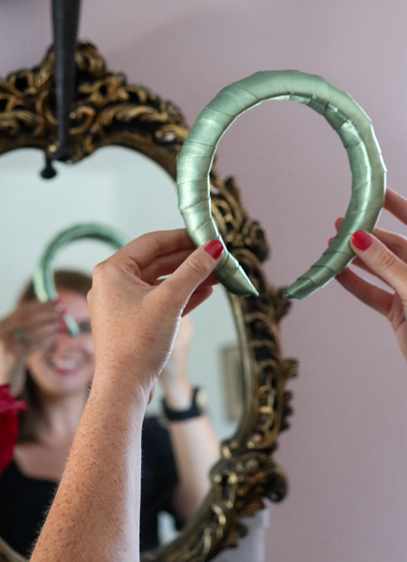 Woman smiling at her reflection while holding up a padded green satin headband in front of an ornate gold mirror, with her nails painted red